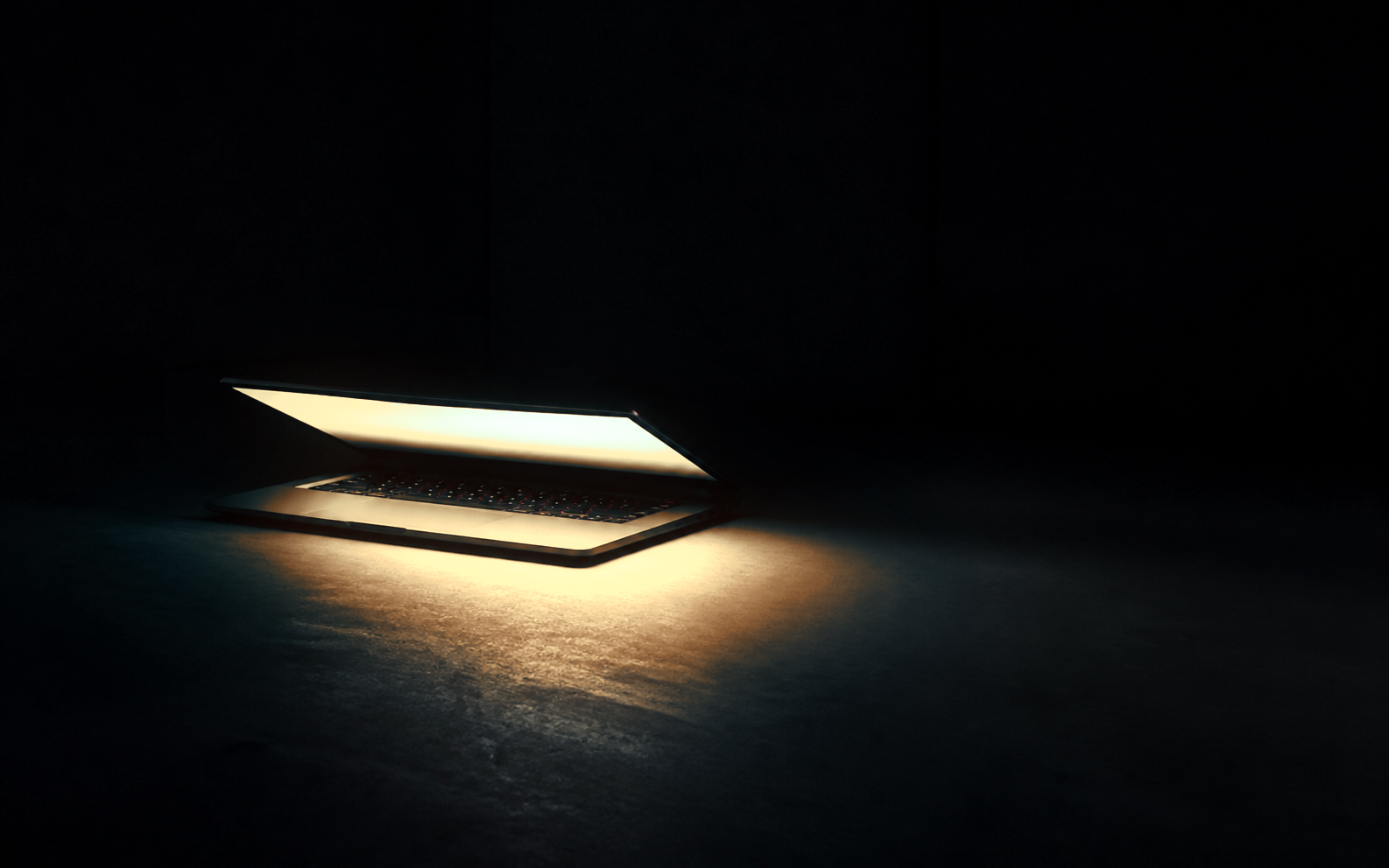 A slightly opened laptop casting warm orange and white light across a dark floor